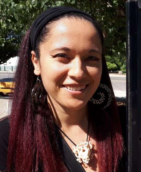 Dr. Deirdre McCorkindale wears a black headband over red dreadlocks posing beside a historical plaque in the outdoors.