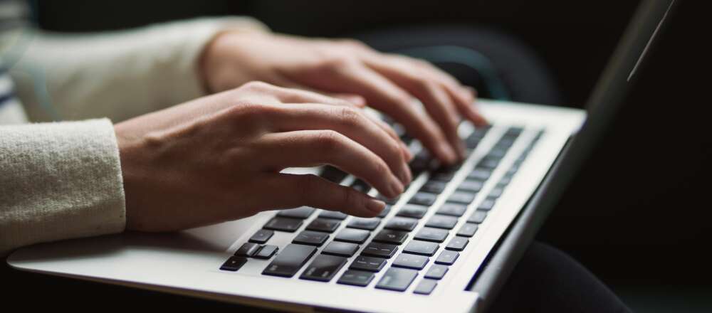 A person wearing a white sweater types on a black keyboard on a pale gray laptop