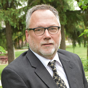 John Zandstra wears a suit and poses for a photo outside in front of evergreen trees