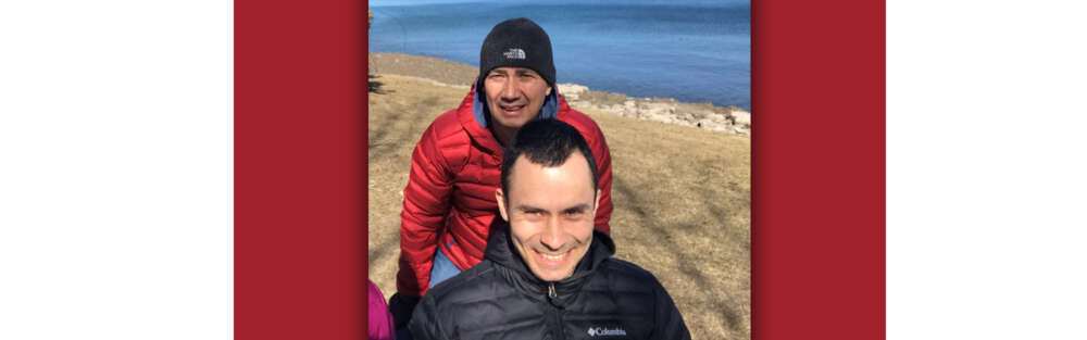 Two men stand on a beach in front of blue water, one standing in a red coat and black toque and the other seated and smiling in a black coat.