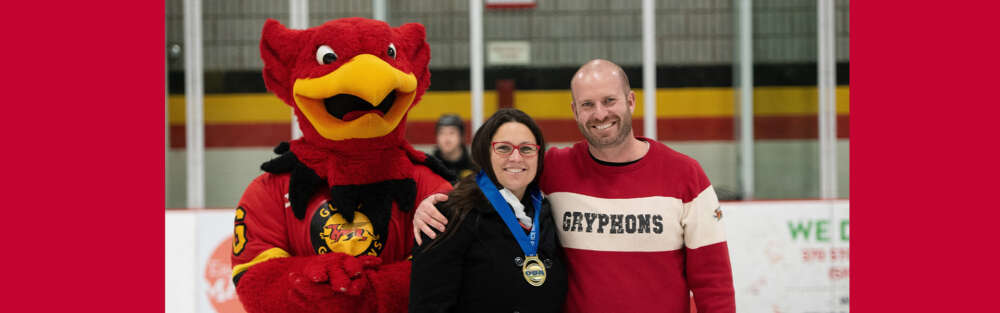 Gryph, the U of G mascot stands beside a person with long brown hair, glasses and a black coat wearing a medal around their neck and a person with a beard and smile wearing a red Gryphons sweater.