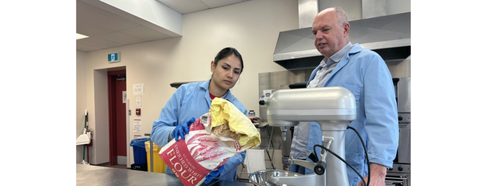 Two people wearing lab coats prepare to pour a bag of flour into a large stand mixer