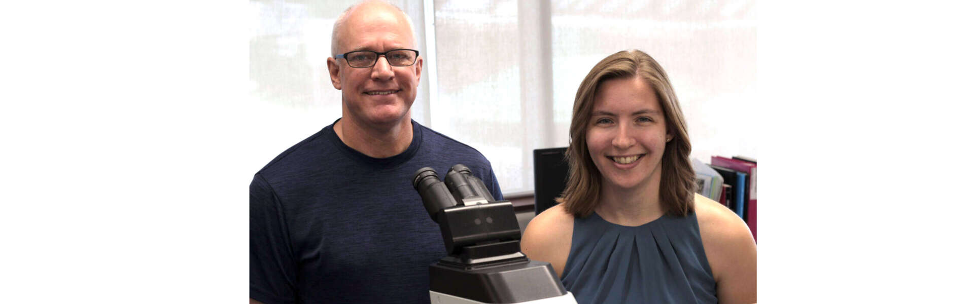 Two people sit near a microscope in a lab, smiling for the camera
