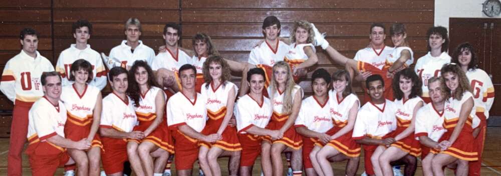 A group of 25 students in cheerleader uniforms pose for a portrait in a university gym
