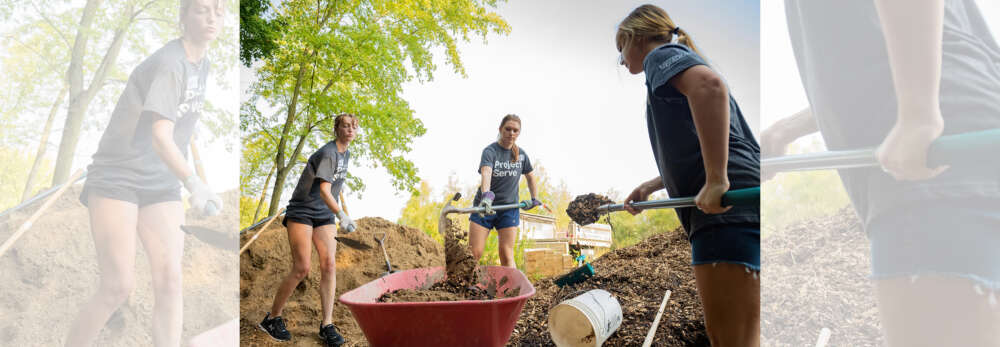 Three students in matching t shirts that say Project Serve shovel dirt into a red wheelbarrow.