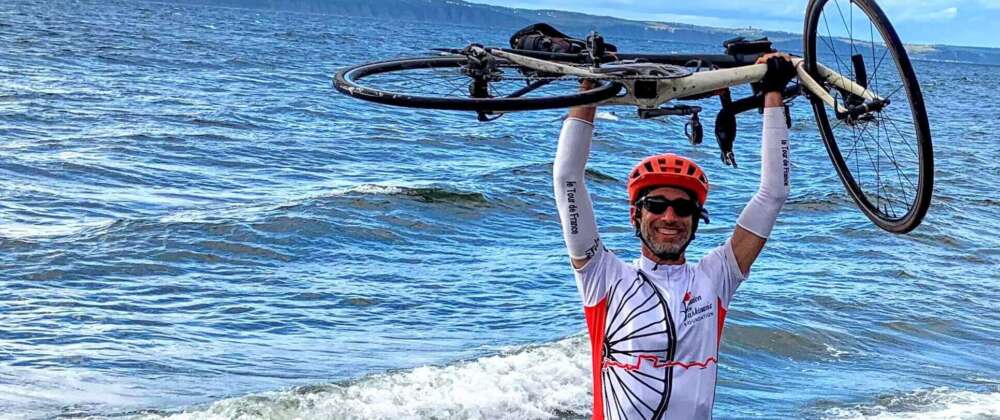 A man in red, white and black cycling clothes raises his bike over his helmeted head standing on the shore of the ocean.