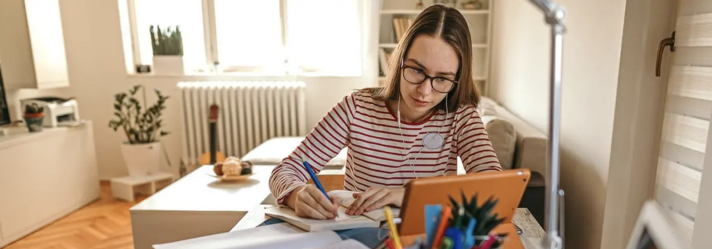 a woman with earbuds sits at a desk in an apartment taking notes with a pen while watching a tablet