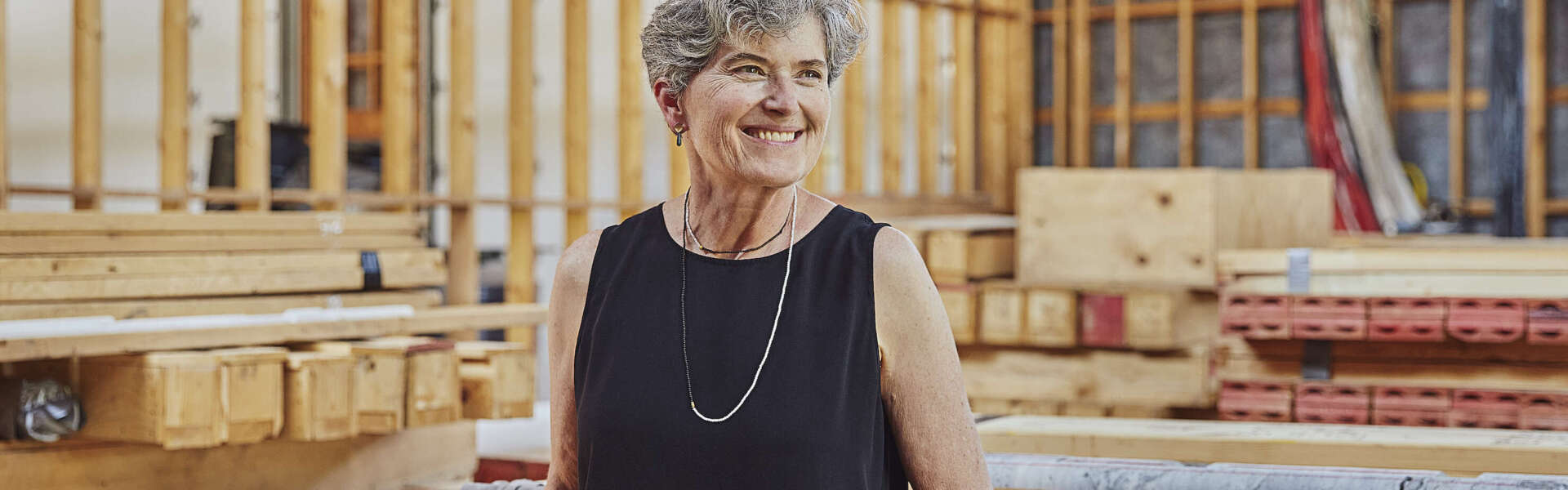 A person with short grey hair wearing a black sleeveless shirt and a silver necklace stands smiling facing left in a workspace with wood behind them.