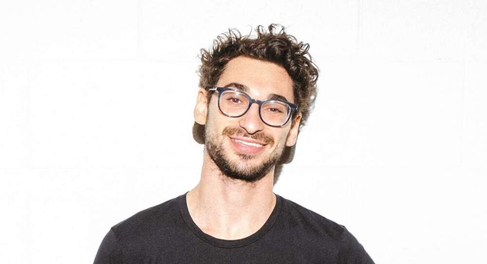 A person with curly black hair, black eyeglasses and black facial hair wearing a black t-shirt smiles standing in front of a white background.