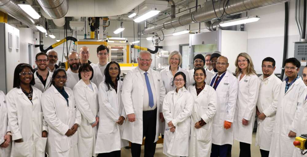 A group of 20 people in lab coats pose for a portrait while standing in a machinery area