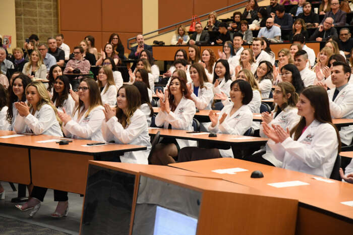 A lecture hall full of OVC students dressed in white coats clap as they sit at desks.