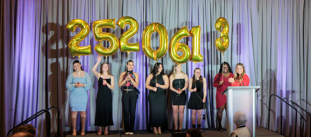 A woman at a podium while seven women in formal wear hold balloons with the numbers 2520613