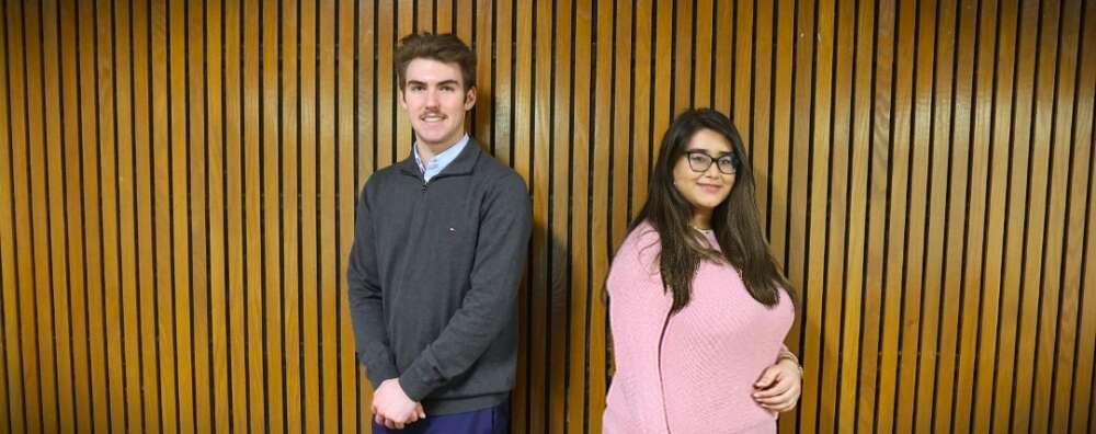 A man and a woman stand back-to-back to pose for a photo against a wood slat wall