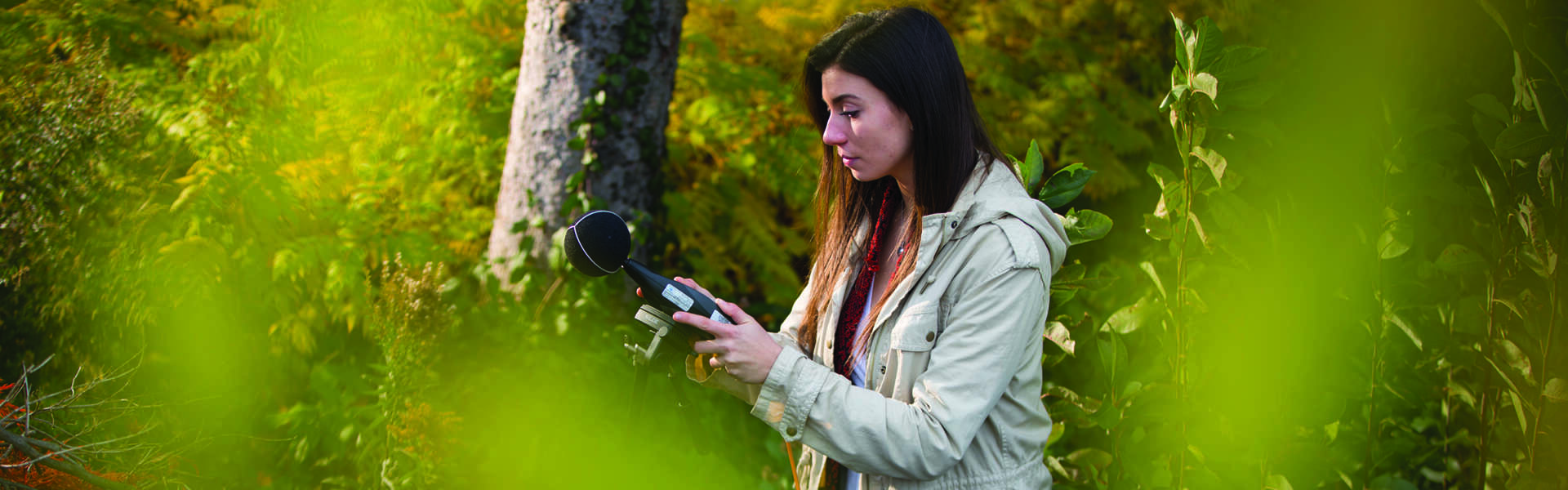 A woman looks at a surveying device while standing in the woods.