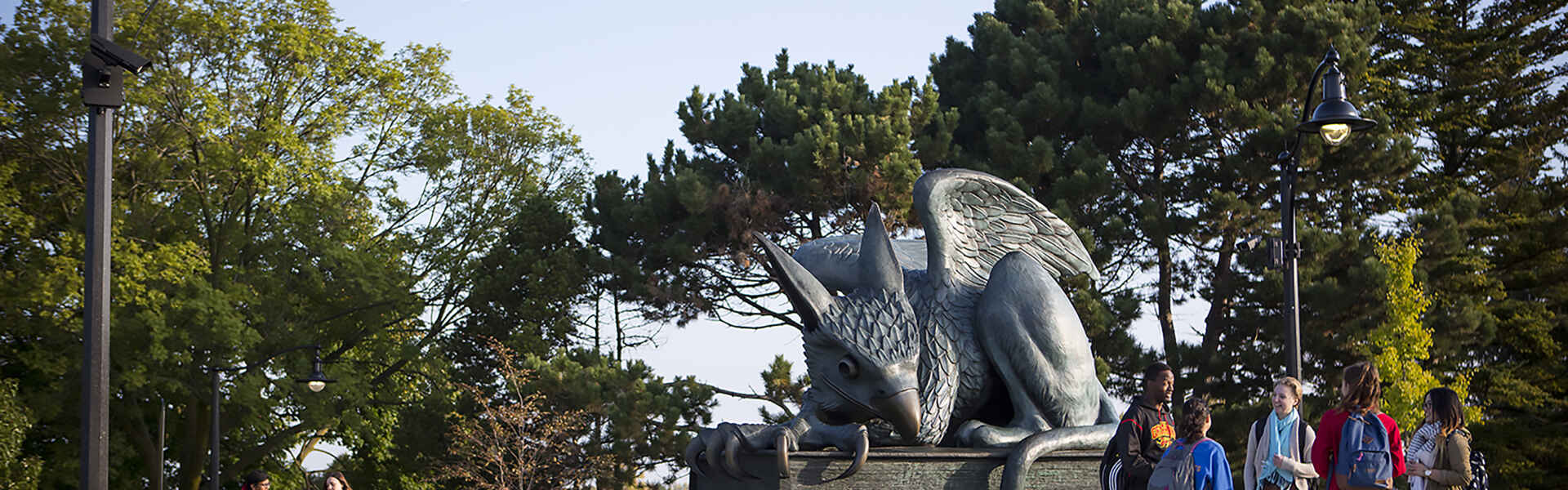 A group of students chat while standing by the Gryphon statue on a sunny day.