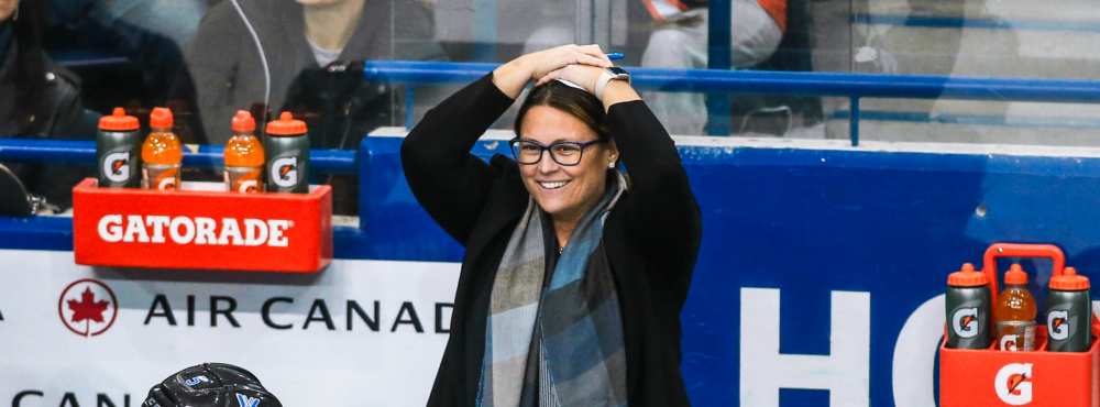A person in a black coat and blue scarf stands with their hands on their head behind a bench full of hockey players in blue uniforms.
