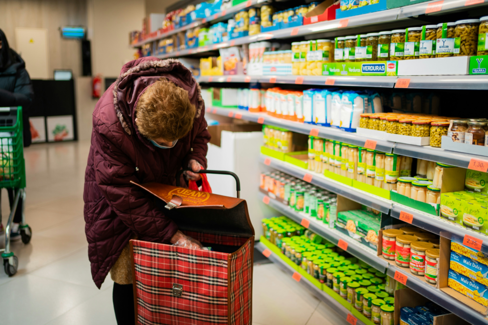 In a grocery store, beside a shelf full of items, a person hunches down to grab something out of their bag.