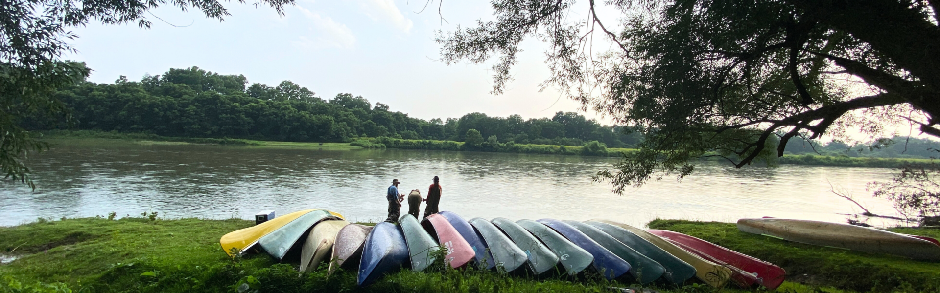 Rows of multi-coloured canoes set on grass, a river and a few canoests stand in the background