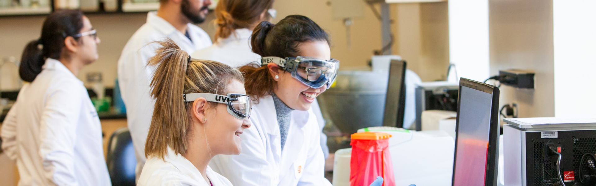 In a classroom laboratory, two students in lab coats, goggles and gloves watch a computer screen, smiling together. Three other students are in the background.