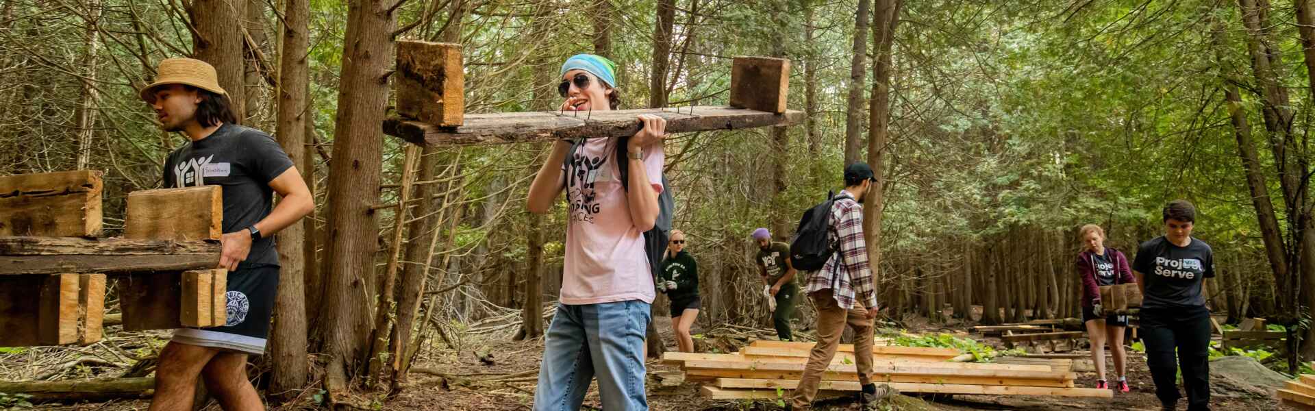 a student in a pink shirt and blue jeans carries a wooden structure through a forest, following a man in a black shirt and shorts