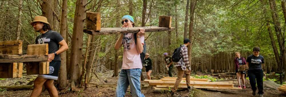 a student in a pink shirt and blue jeans carries a wooden structure through a forest, following a man in a black shirt and shorts