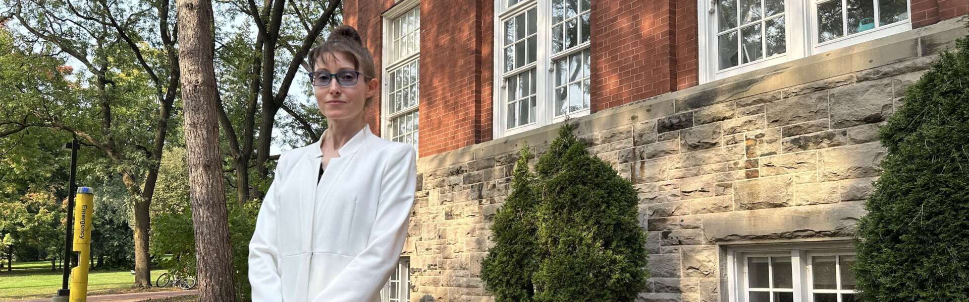 A person in a white overcoat, brown hair and glasses stands in front of a red brick building with trees surrounding.