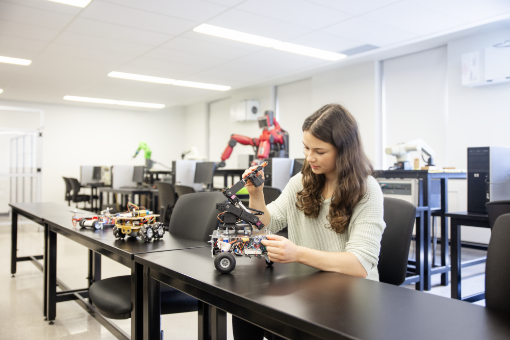 A student is seated in a lab surrounded by computers and robotic equipment. She is working on a wheeled robot with a robotic arm. Other robots and machinery are visible in the background, creating a modern robotics lab atmosphere.