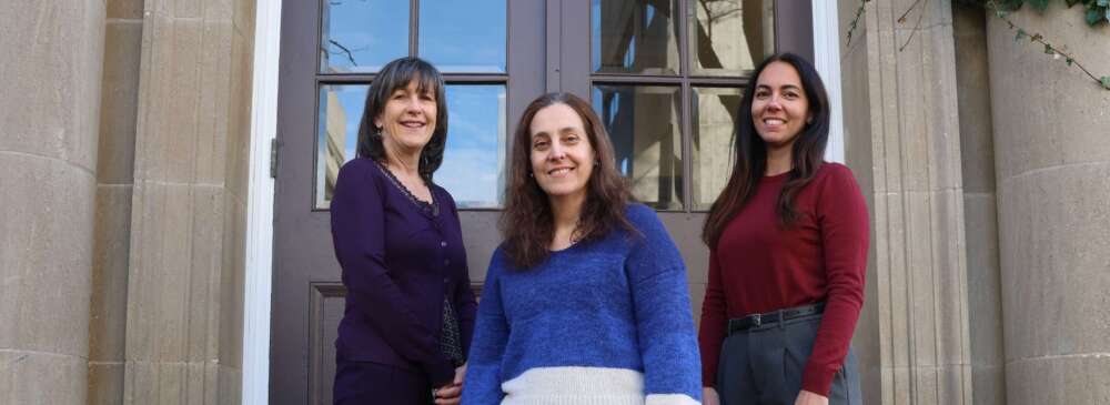 Three people with brown hair wearing purple, blue and red sweaters stand on the steps of a brick building in front of a wooden door with a glass window.