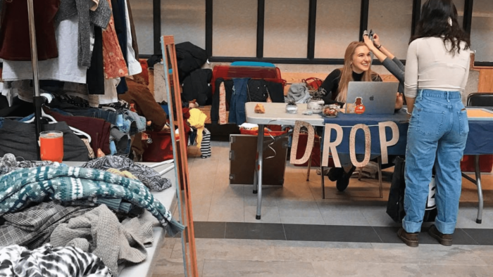 Two women talk at a table labeled drop. To the left is a collection of clothes.