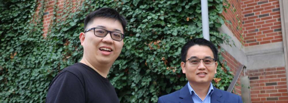 Two people with glasses, on wearing a black t-shirt and one wearing a blue button-up under a blue blazer stand in front of greenery and a red brick wall.