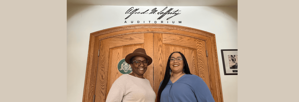 Two Black women stand facing one another and smiling, one in a beige sweater and brown hat and one in a blue shirt and black braids in front of two wooden doors with script on the wall above them.