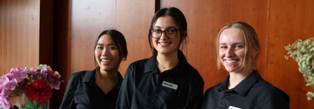Three students in black button-up long sleeve shirts stand together smiling behind a counter with a vase of multicoloured flowers in front of a wooden wall.