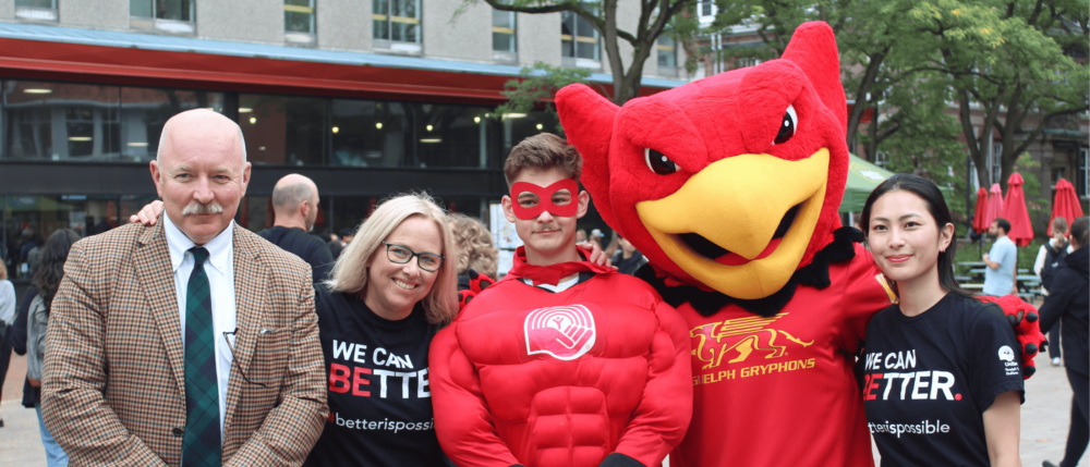 A group of five volunteers for the United Way campaign. Two wear "We can better" shirts, one wears a red superhero costume and one wears a Guelph Gryphons costume.