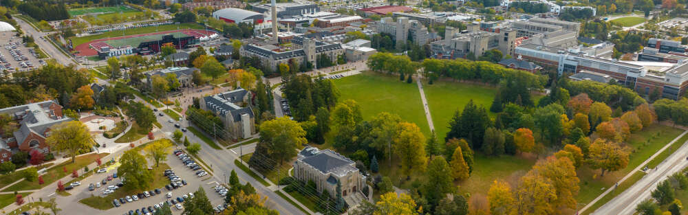 An aerial view of the University of Guelph campus depicts multiple buildings, Johnston green, the football stadium and lush greenery.