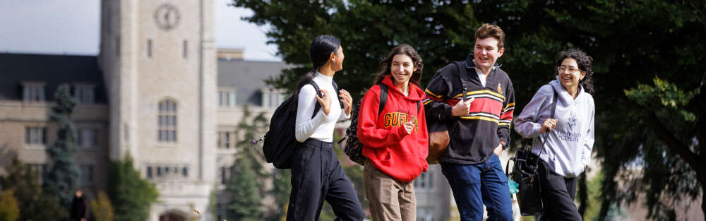 A group of four students talk and laugh as they walk across the U of G campus with Johnston Hall and mature trees in the background.