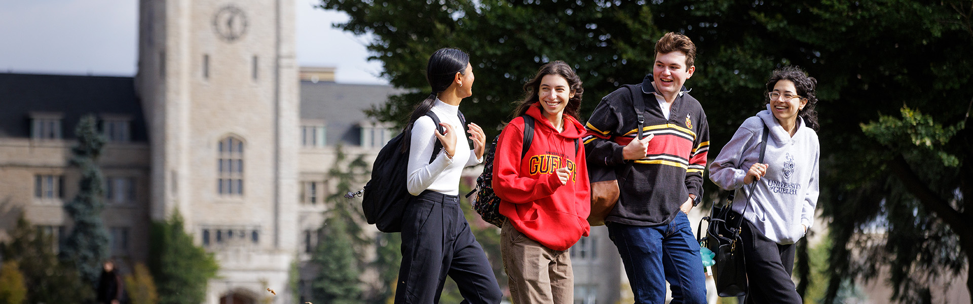 A group of four students talk and laugh as they walk across the U of G campus with Johnston Hall and mature trees in the background.