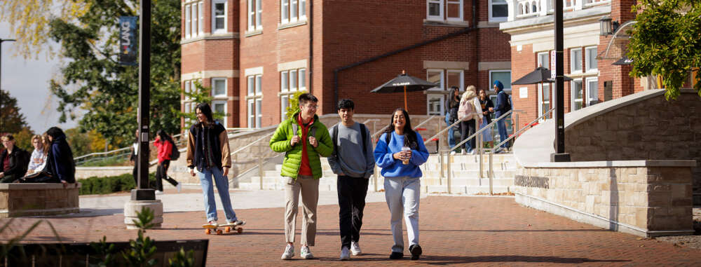 Three students walk together smiling in front of Macdonald Hall while groups of students gather on the steps behind them and another skateboards on the cobblestone.