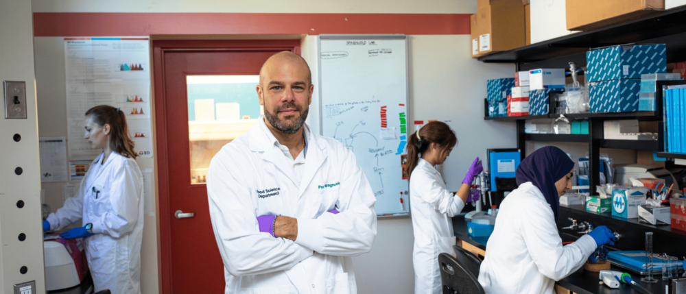 Dr. Paul Spagnuolo smiles at the camera as a few researchers work behind them in a lab