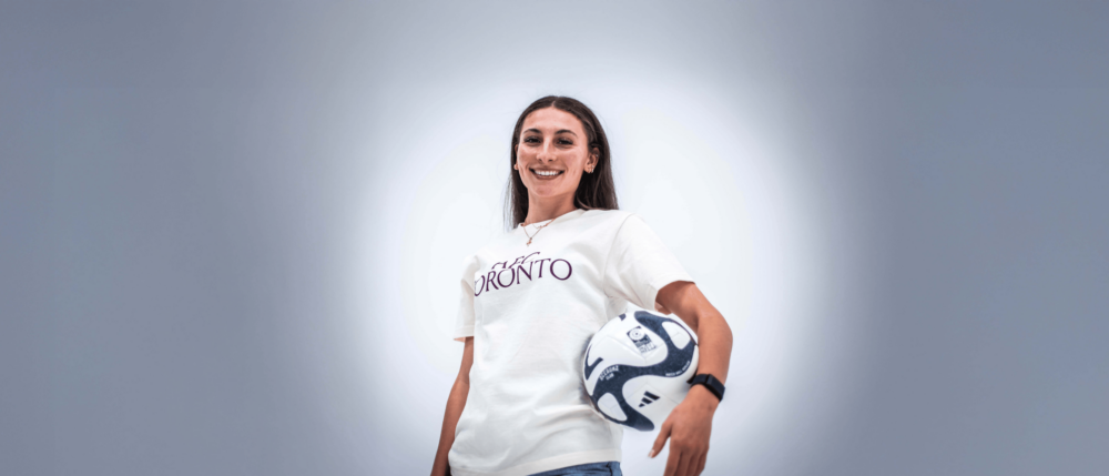 A woman with long brown hair stands smiling with a blue and white soccer ball under one arm while wearing a white t-shirt with a maroon AFC Toronto logo on the front.