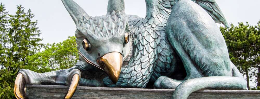 A close-up of a large, intricately designed griffin statue made of metal with a weathered, greenish patina. The mythical creature, a hybrid of an eagle and a lion, has a sharp, golden beak and piercing golden eyes. Its head is adorned with textured, overlapping feathers, while its powerful claws grip the edge of a surface.
