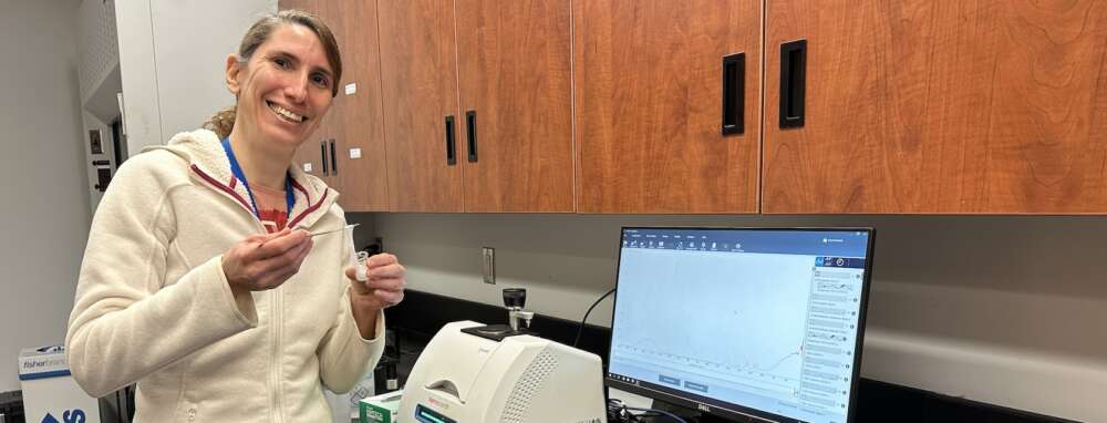 A person wearing a cream-coloured zip-up jacket stands smiling into the camera while holding a small glass jar and tool to display a clear-coloured substance inside, standing in front of brown cabinets and a computer screen and printer.