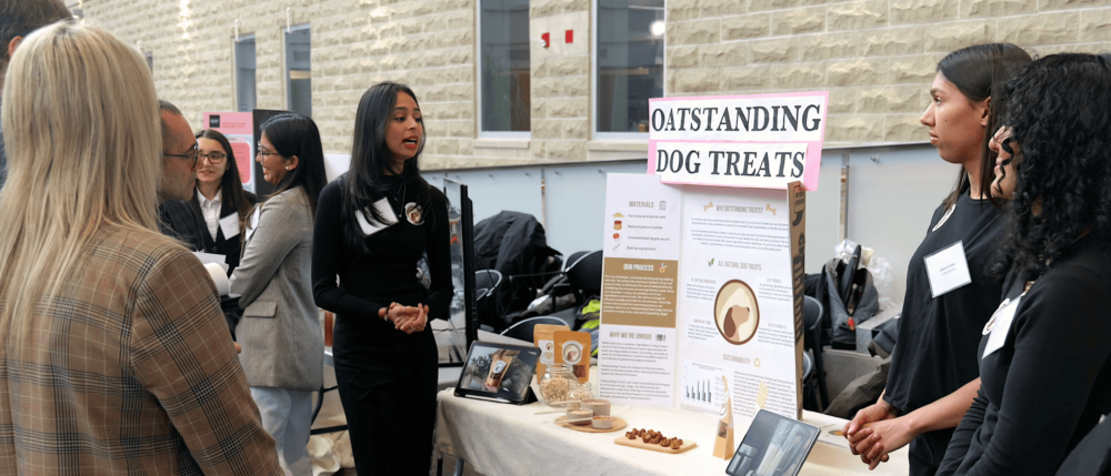 Student presenter stands in front of a display entiteld 'Oatstanding Dog Treats' as observers watch. Display contains food samples of their project
