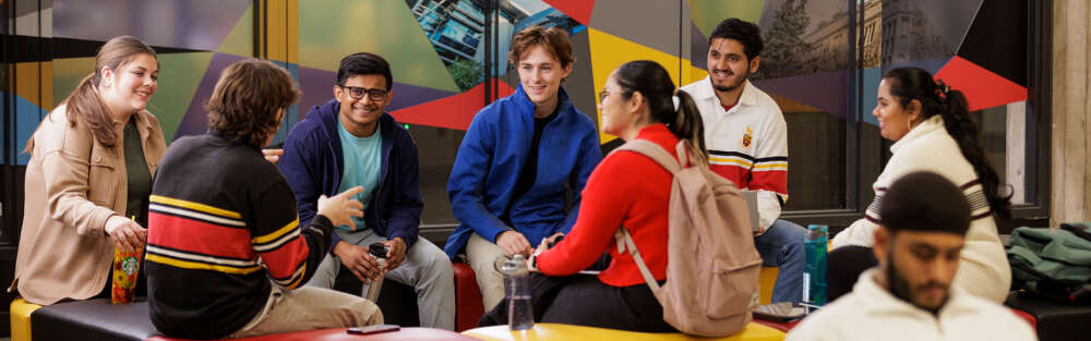 A group of seven diverse students sit in a brightly coloured lounge--U of G red, black and gold colours prominent--conversing with one another.