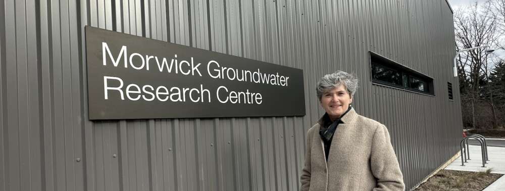 A person with short, curly grey hair wearing a purple scarf and beige coat stands with their hands in their pockets in front of a dark grey building with a black sign with white text that reads Morwick Groundwater Research Centre.