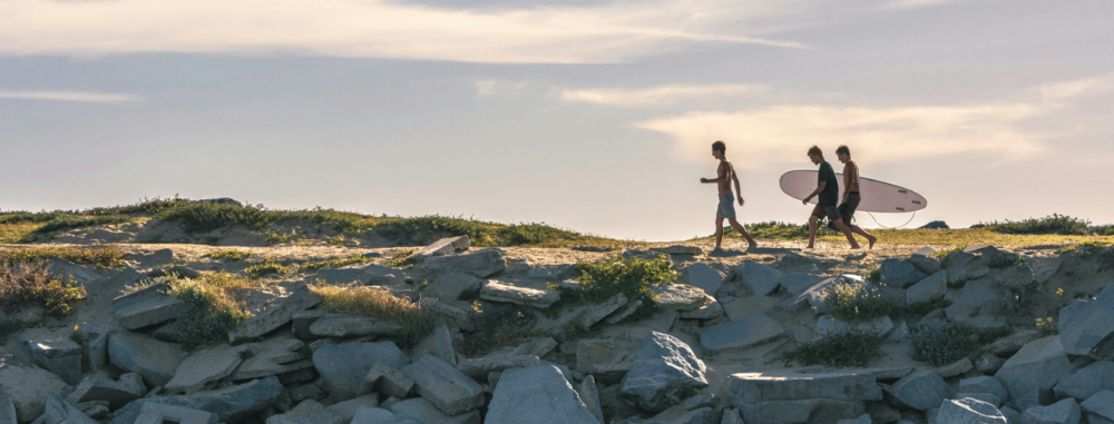 Three people walk along a rocky shoreline under a clear sky, with two of them carrying surfboards, heading toward the ocean. The sun casts a warm glow over the scene, highlighting the texture of the rocks and the gentle vegetation.