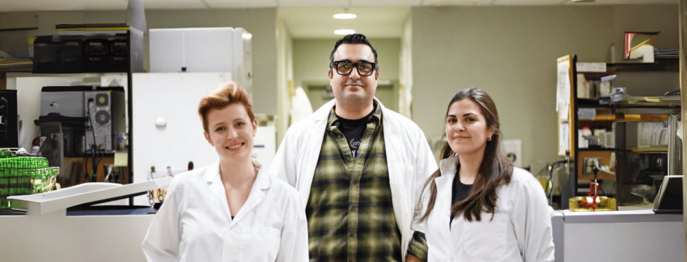 Three researchers wearing white lab coats stand together in a laboratory setting, smiling at the camera. The researcher on the left has short, styled hair and is wearing purple gloves. The researcher in the middle wears glasses and a plaid shirt under their lab coat. The researcher on the right has long brown hair and a black shirt under their lab coat. Laboratory equipment and shelves are visible in the background.