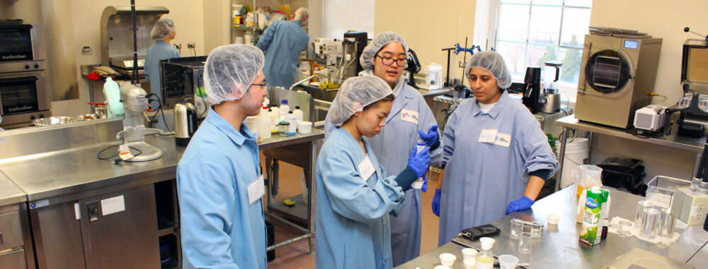 four students dressed in blue lab coats and white hair nets look at food products on a bench in a lab