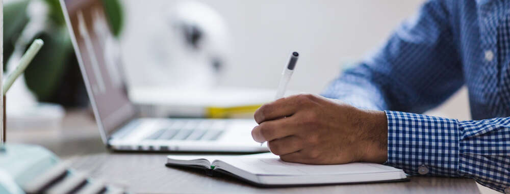 a hand writes in a notebook, an open laptop in the background