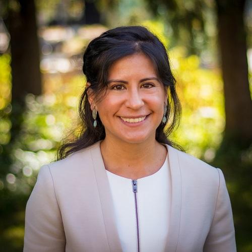 a woman with dark hair wearing a white shirt and beige jacket poses for a headshot