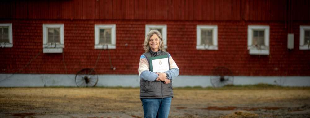 a woman in a striped shirt, grey vest and blue jeans poses for a photo holding a certificate in a folio, with a red barn in the background